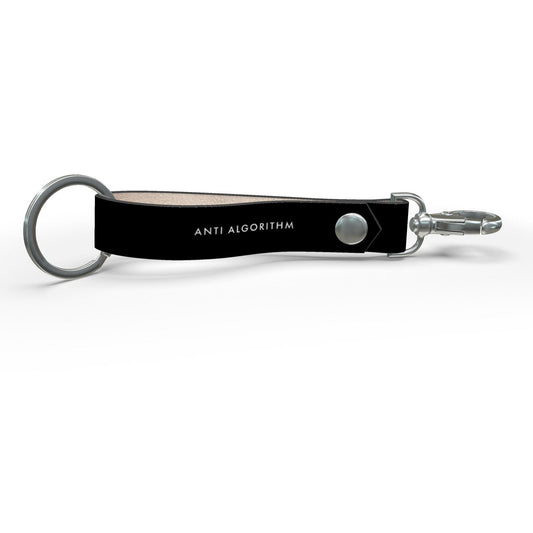 Side view of a black leather key strap with white ANTI ALGORITHM lettering, a silver-tone ring and clip